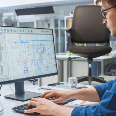 Over the Shoulder Shot of Engineer Working with CAD Software on Desktop Computer, Screen Shows Technical Drafts and Drawings. In the Background Engineering Facility Specialising on Industrial Design
